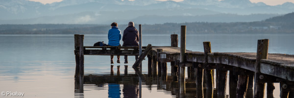 Ein Paar sitzt auf einem Bootssteg am Ammersee und sieht auf das Berg-Panorama